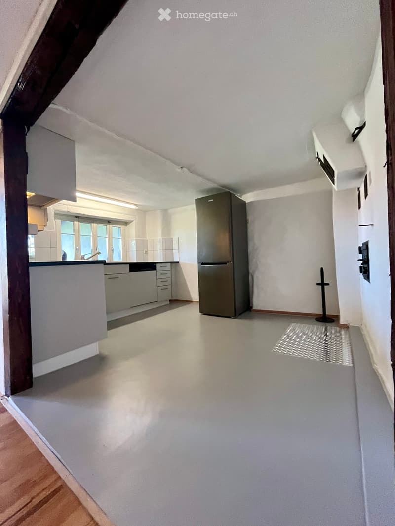 Empty room with white walls, light-colored floor, and a small kitchen area visible in the background. No furniture or appliances are present.