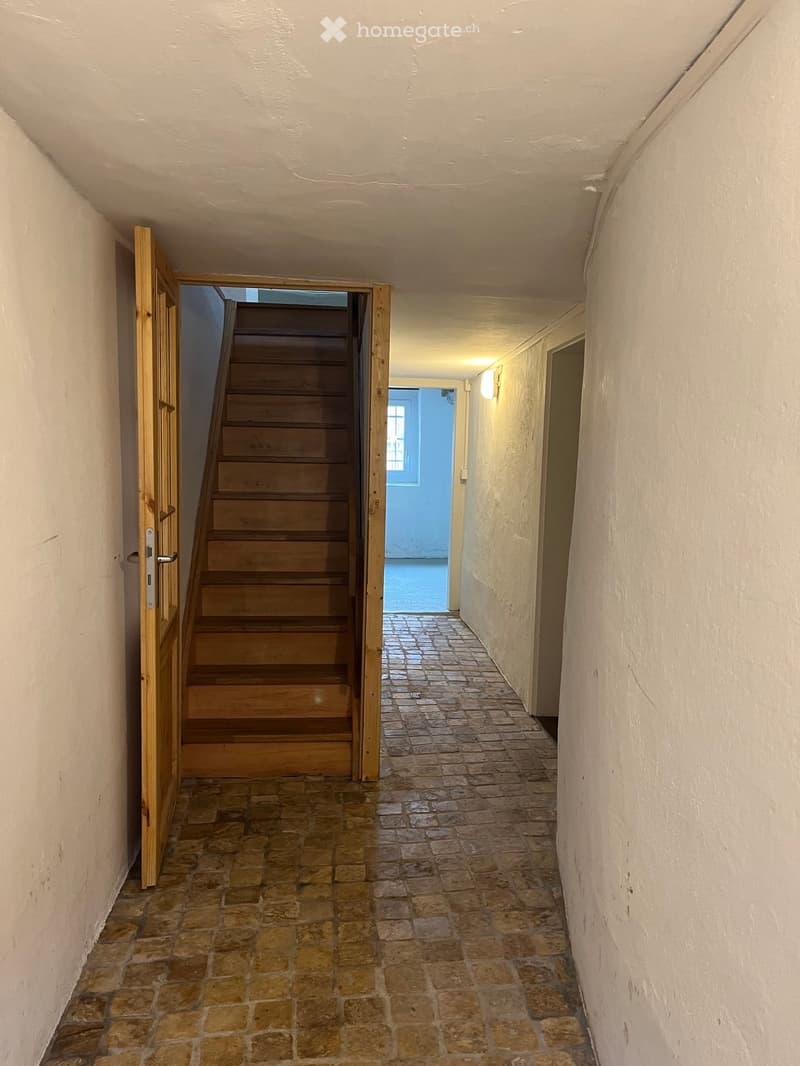 A narrow hallway with a brick floor, white walls, and a wooden staircase leading upwards. A wooden door is open on the left, and a light fixture is mounted on the right wall.