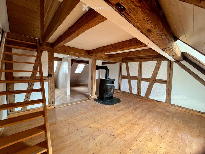 An attic space with wooden beams, wooden flooring, and a staircase leading to another area. A wood-burning stove is present, and there are skylights providing natural light.
