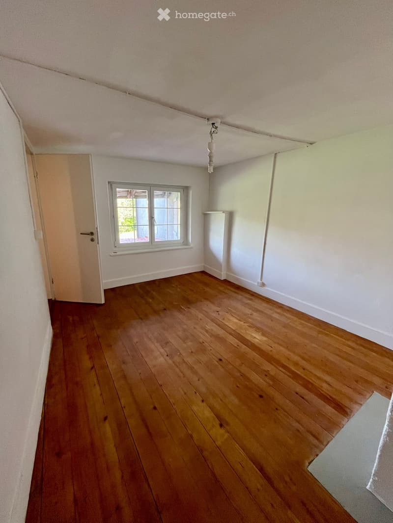Empty room with wooden flooring, white walls, a single window, and a door leading to another space.