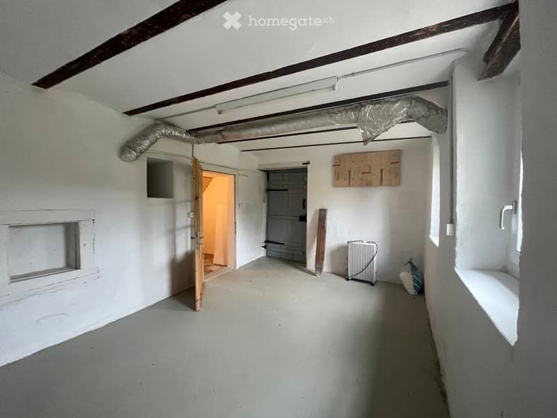 Empty room with white walls, exposed wooden beams, and a concrete floor. Features include a fireplace niche, a doorway leading to another room, and a small radiator. The space appears unfinished with minimal furnishings.