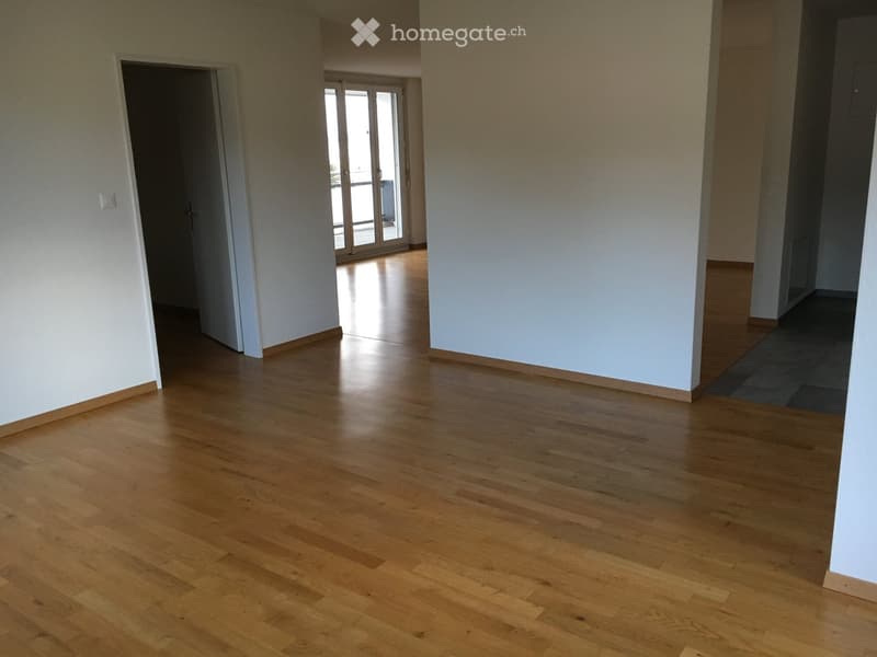 Empty room with light wooden flooring and white walls, featuring an open doorway leading to another room with large glass doors and a view of the outside.