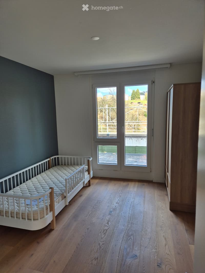A small, unfurnished room with wooden flooring, a single bed, and a wardrobe. The room has large windows with a view of the outside.
