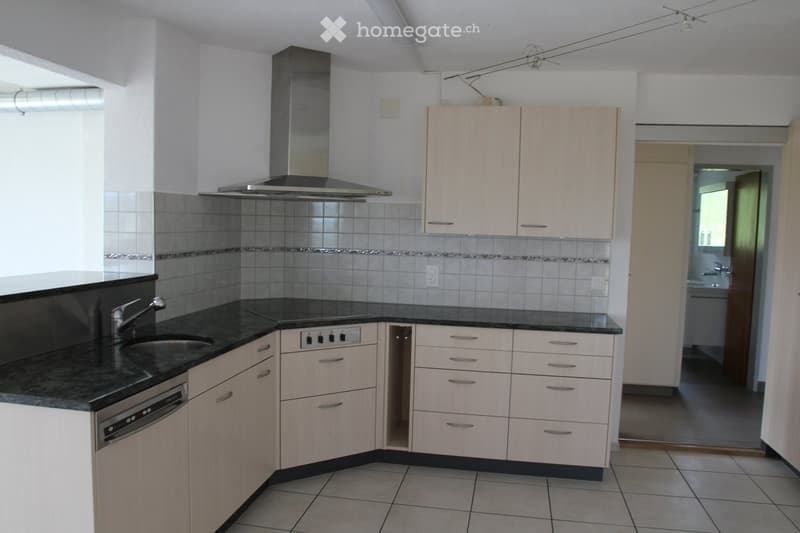 Modern kitchen with light wood cabinets, granite countertops, stainless steel sink, dishwasher, and range hood. The space is clean and well-organized, with tiled flooring and a visible bathroom area in the background.