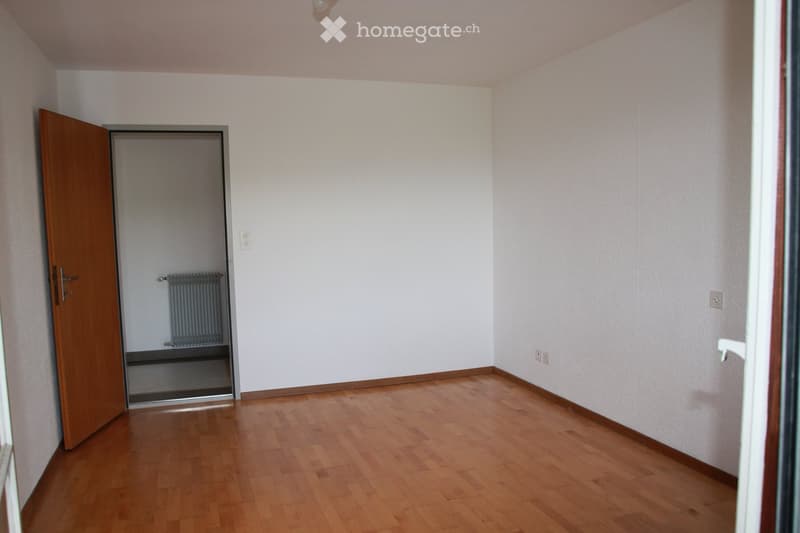 Empty room with light-colored walls, wooden flooring, and a partially open wooden door leading to another room with a radiator visible.