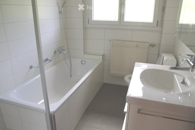 Modern bathroom with white tiled walls, a bathtub, a sink with a mirror, a toilet, and a radiator.