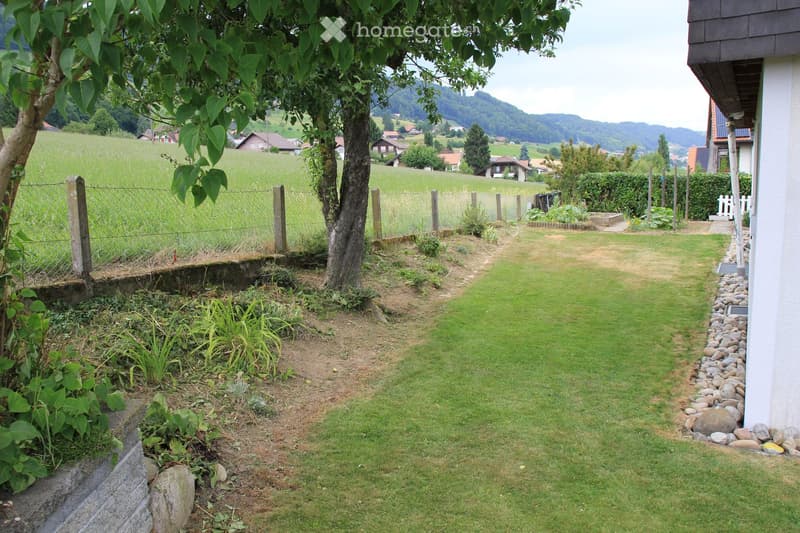 A well-maintained garden with a grassy lawn, a stone border, a small garden bed with plants, and a fence separating the garden from a field.