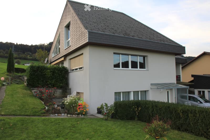 Two-story house with a modern design, grey roof, white walls, multiple windows, and a well-maintained garden with various plants and shrubs.