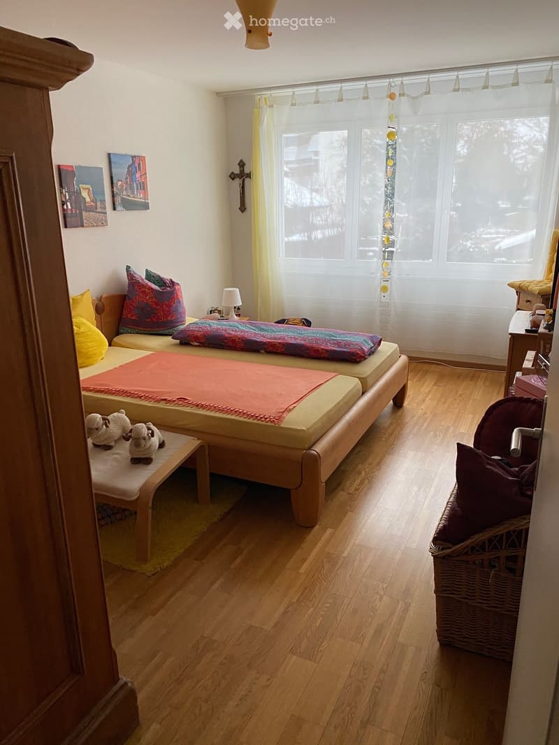 Cozy bedroom with wooden flooring, a bed with colorful blankets, a small sofa, a wooden stool, a table, a wicker basket, and decorative items including a cross and picture frames.