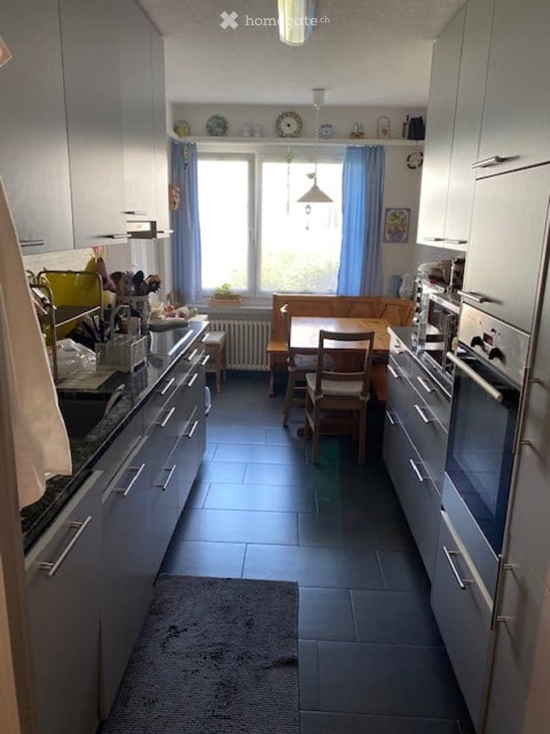 Modern kitchen with grey cabinets, a central island, and a dining area with a table and chairs. The space features a window with blue curtains, a sink, and various appliances including an oven and microwave.