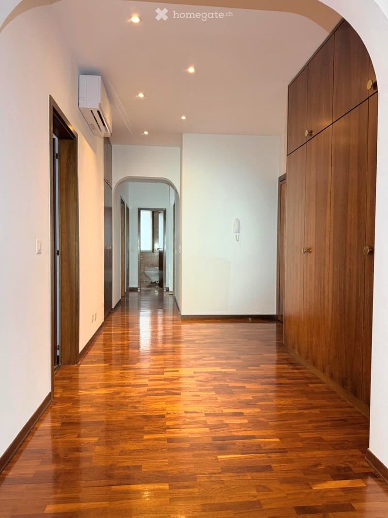 Spacious hallway with polished wooden flooring, white walls, and recessed lighting. Features built-in wooden cabinetry on one side and an open doorway leading to another room.