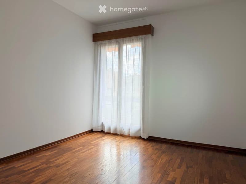 Empty room with wooden flooring, white walls, and a window with sheer curtains.