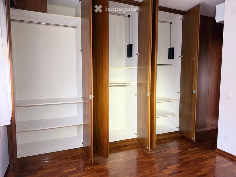 Empty walk-in closet with wooden doors, white walls, and wooden flooring. The closet features multiple shelves and hanging rods, but no items are present.