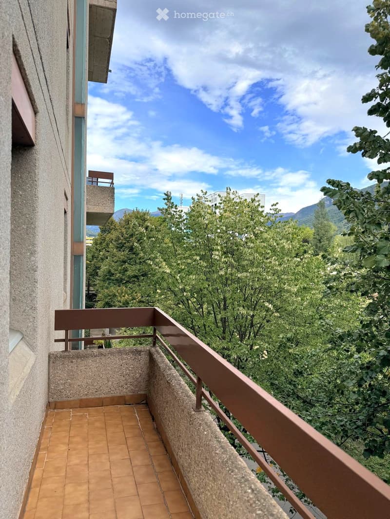 Balcony with tiled floor, metal railing, overlooking a green, tree-filled area with mountains in the background.