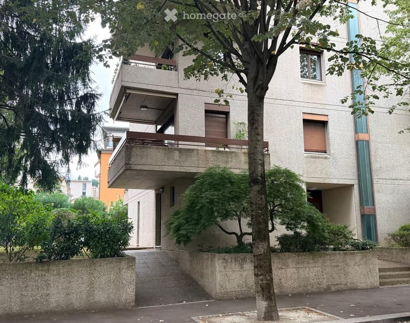 Modern multi-story building with balconies, large windows, and surrounding greenery.