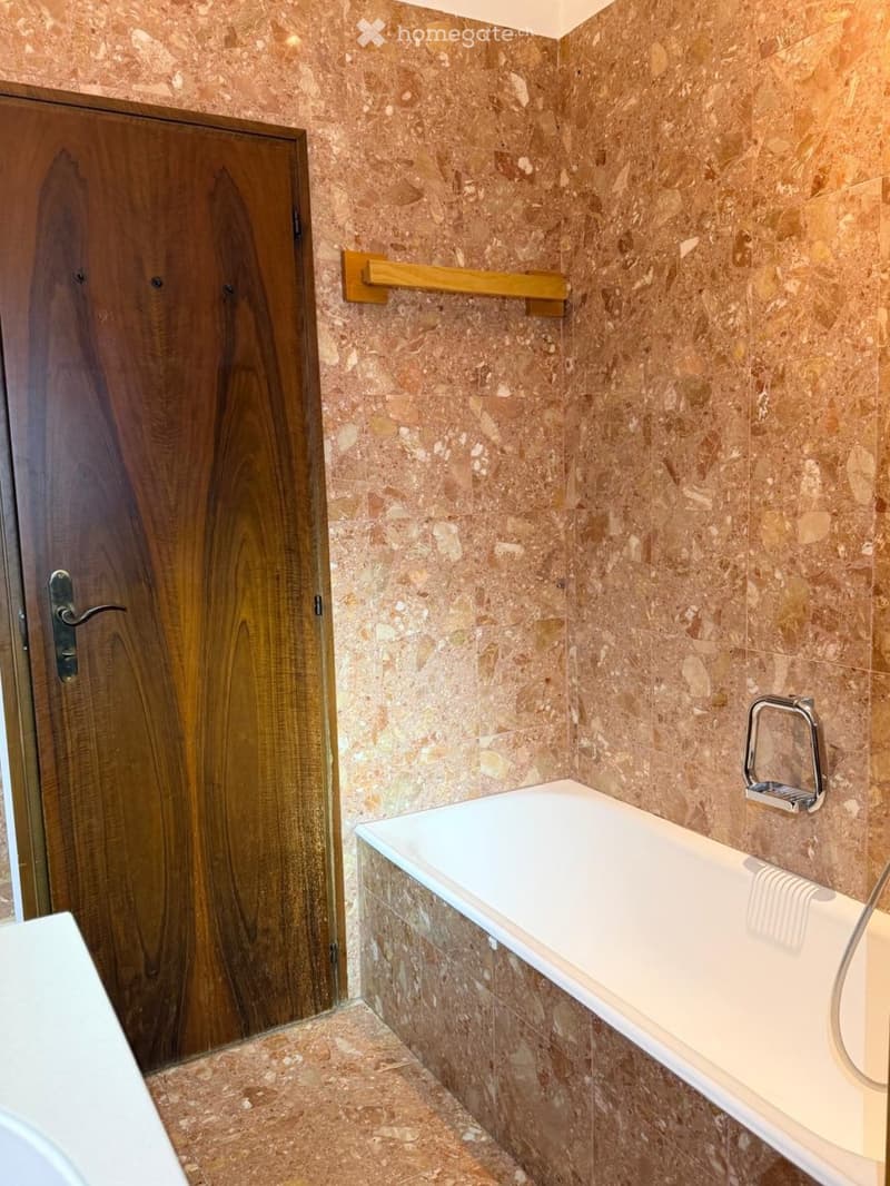 Bathroom with marble walls and floor, a bathtub, a towel rack, and a wooden door.