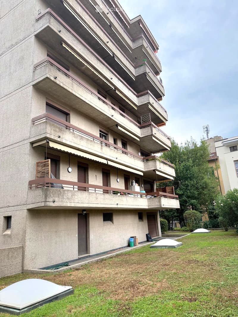 Multi-story residential building with balconies, concrete facade, and surrounding greenery.