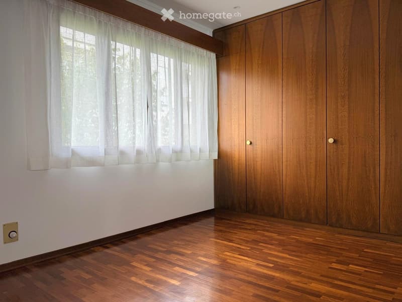 Empty room with wooden flooring, white walls, large window with white curtains, and wooden closet doors.
