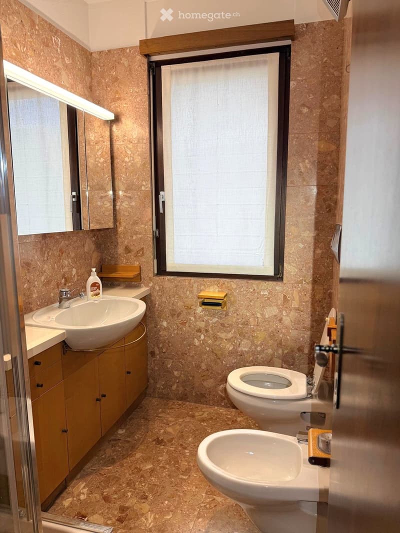 Bathroom with beige marble tiles, a sink with a modern faucet, a toilet, a bidet, and a mirrored cabinet. A window with blinds provides natural light.