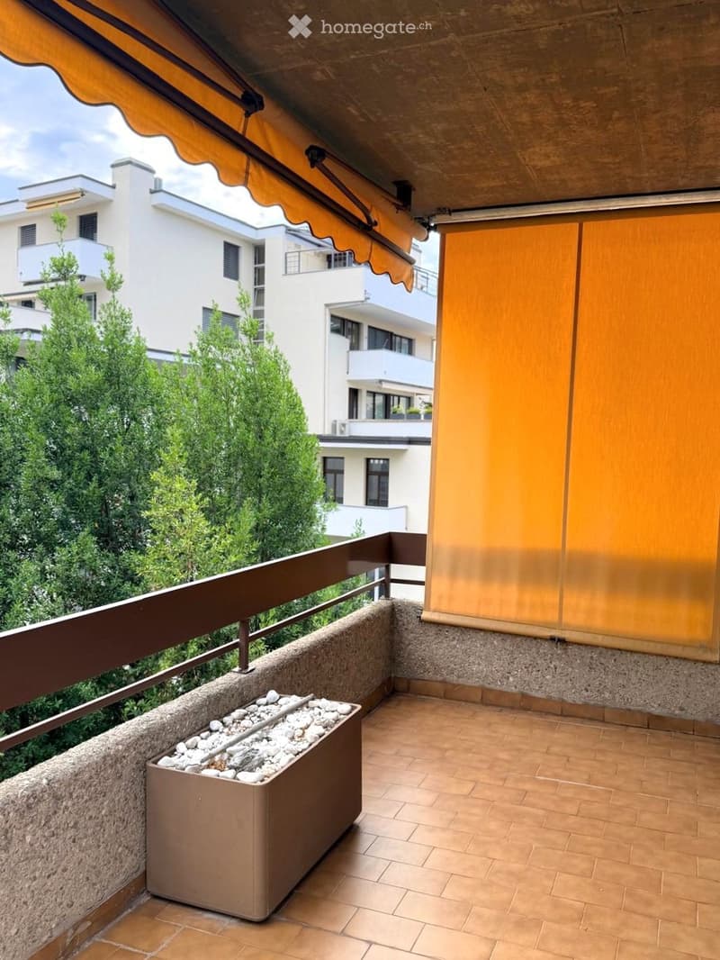 Balcony with tiled floor, brown railing, and a planter with white pebbles. Features an orange awning and a view of neighboring buildings and greenery.