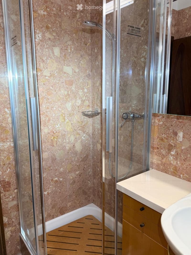 Modern bathroom with marble walls, a glass-enclosed shower, a wooden shower floor, a sink with a modern faucet, and a small shower basket.