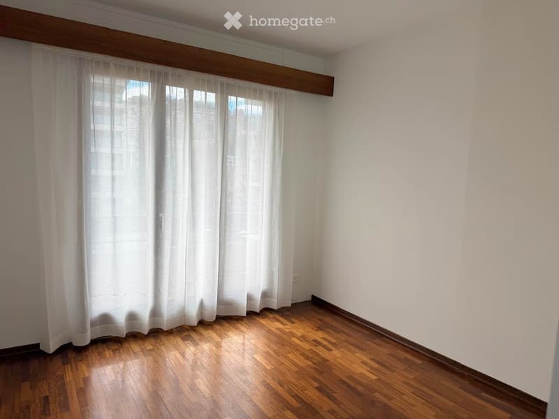 Empty room with wooden flooring, white walls, and large windows covered by white curtains.