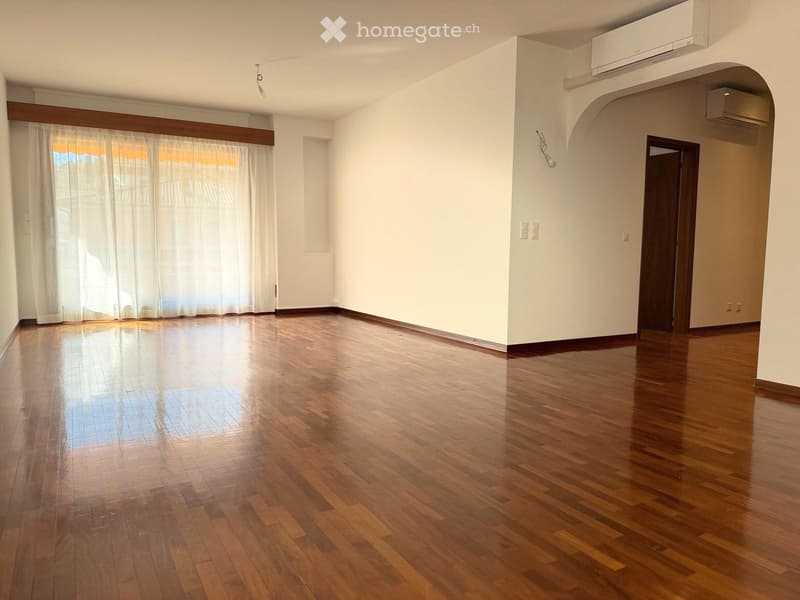Empty room with wooden flooring, white walls, large window with white curtains, and an arched doorway leading to another room.