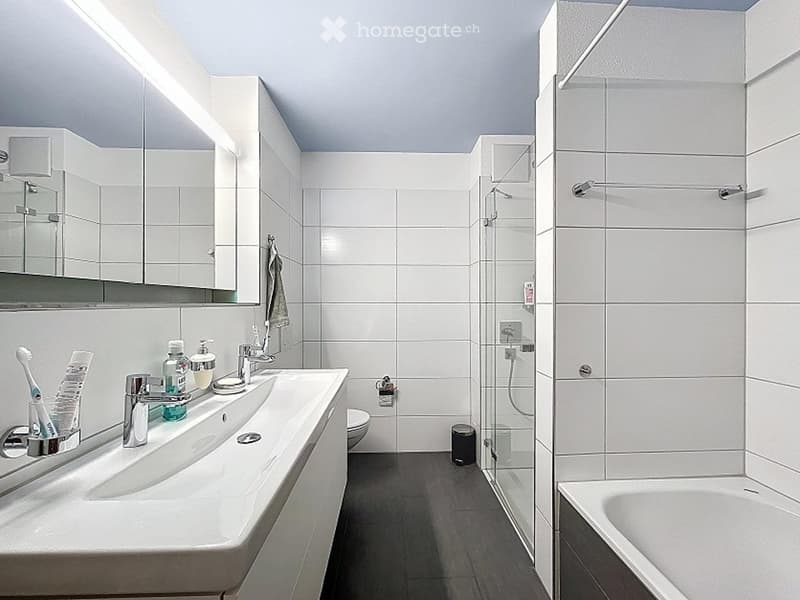Modern bathroom with white tiles, a large sink with a mirror cabinet, a bathtub, and a glass-enclosed shower. The sink has a toothbrush holder, toothpaste, and soap dispenser. A towel hangs on a rack, and a small trash can is placed near the toilet.