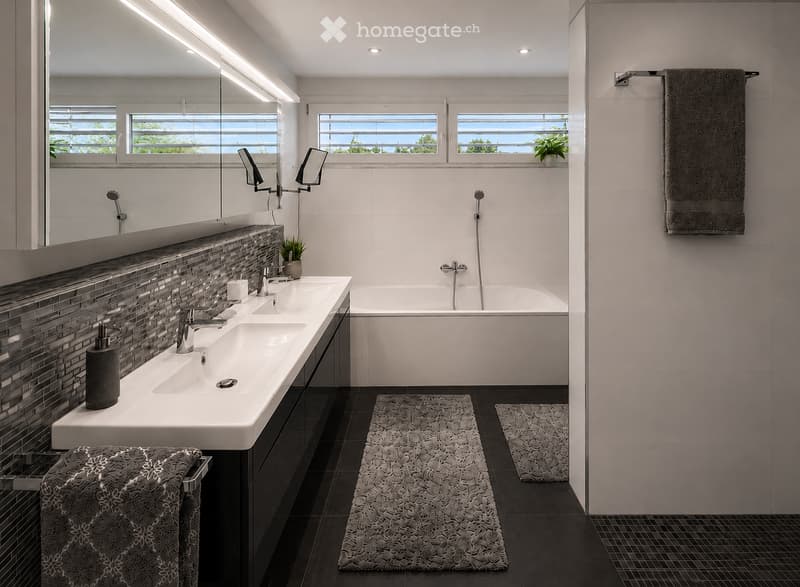 Modern bathroom with double sink, bathtub, large mirror, towel, soap dispenser, and small plant on windowsill.