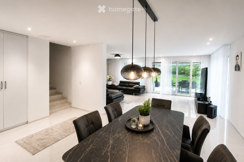Modern, open-concept living and dining area with white walls, polished tile flooring, and large windows leading to a patio. Features a dark wood dining table with six chairs, a black leather sofa and ottoman, a television on a stand, and a pendant light f