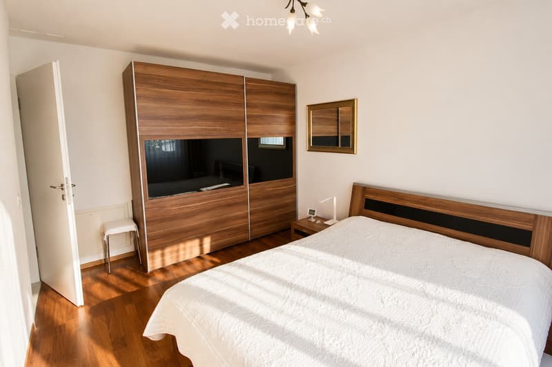 Modern bedroom with a large wooden bed, nightstand, mirror, wardrobe, and a small stool near the door.