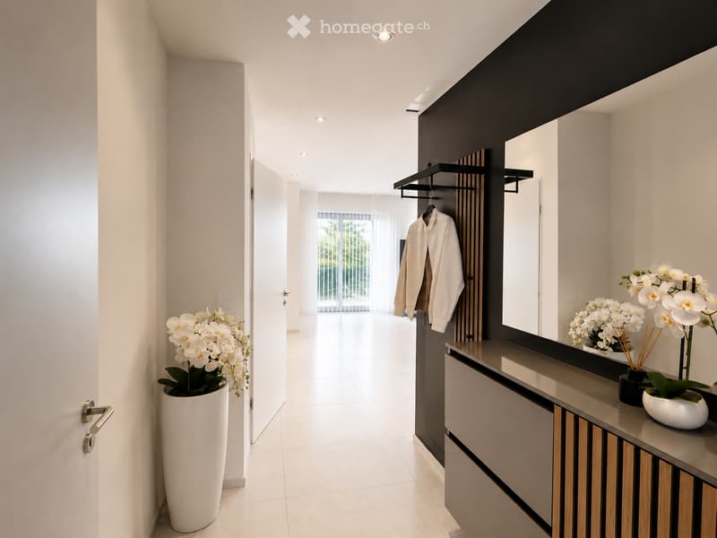 Modern hallway with white walls, tiled floor, recessed lighting, and a large mirror. Features a coat rack with a white jacket, a tall white vase with orchids, and a sleek console table with decorative items.