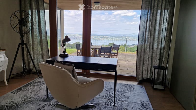 Bright room with large windows offering a scenic view, furnished with a comfortable chair, a desk, a lamp, and a fan.