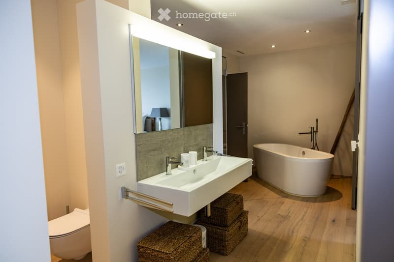 Modern bathroom with a white sink, bathtub, toilet, and large mirror with built-in lighting. Wicker baskets are placed under the sink.