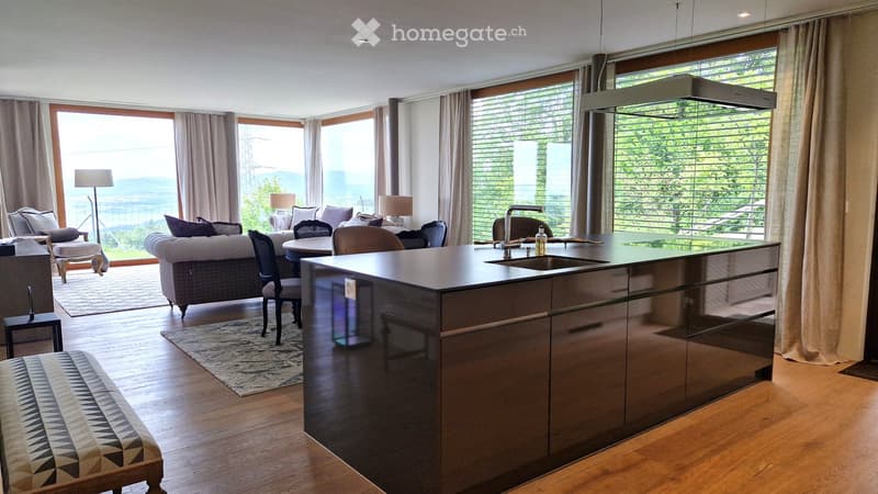Modern open-plan living and dining area with large windows offering a scenic view, featuring a sofa, chairs, a dining table, a kitchen island with a sink, and decorative lamps.