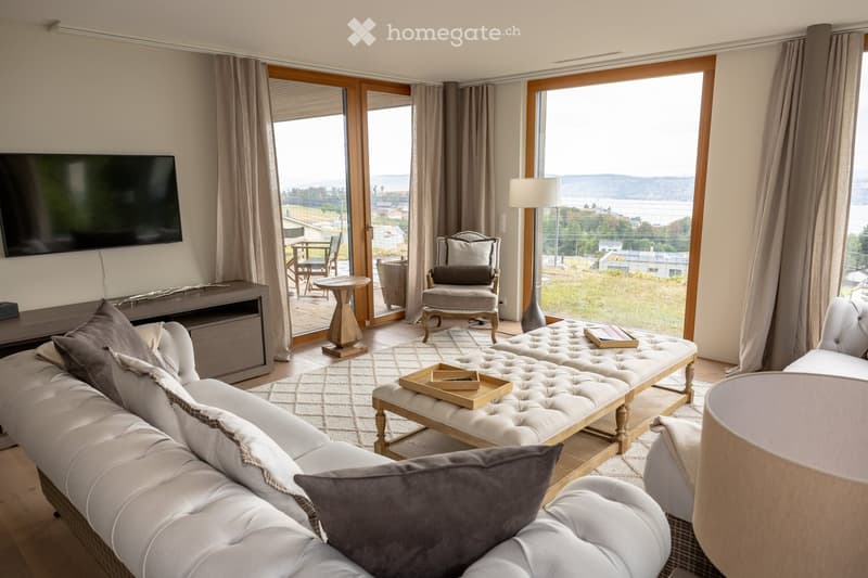 Modern living room with large windows offering a scenic view, featuring a white tufted sofa, coffee table, armchair, floor lamp, and a television mounted on the wall.