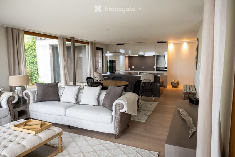 Modern living room with a white sectional sofa, wooden coffee table, and a dining area with a round table and chairs. The space features large windows with curtains, wooden flooring, and a sleek kitchen area in the background.