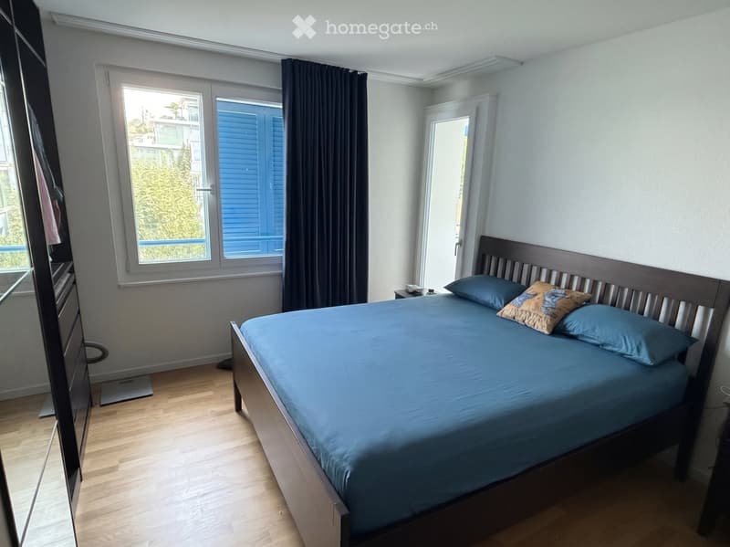 Bedroom with a wooden bed, blue bedding, nightstand, and wardrobe. Large windows with dark curtains and a view of greenery.