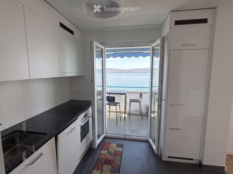 Modern kitchen with white cabinets, black countertops, and a view of the sea through large glass doors leading to a balcony.