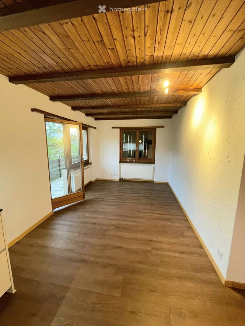 Empty room with wooden floor, exposed wooden ceiling beams, and a large window with a view of the balcony.