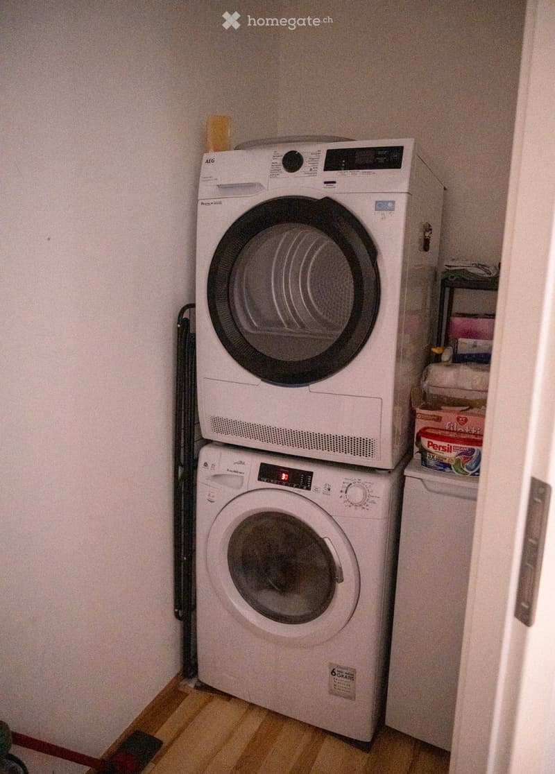 A compact bathroom with a stacked washing machine and dryer unit, wooden flooring, and a small shelf with cleaning supplies.