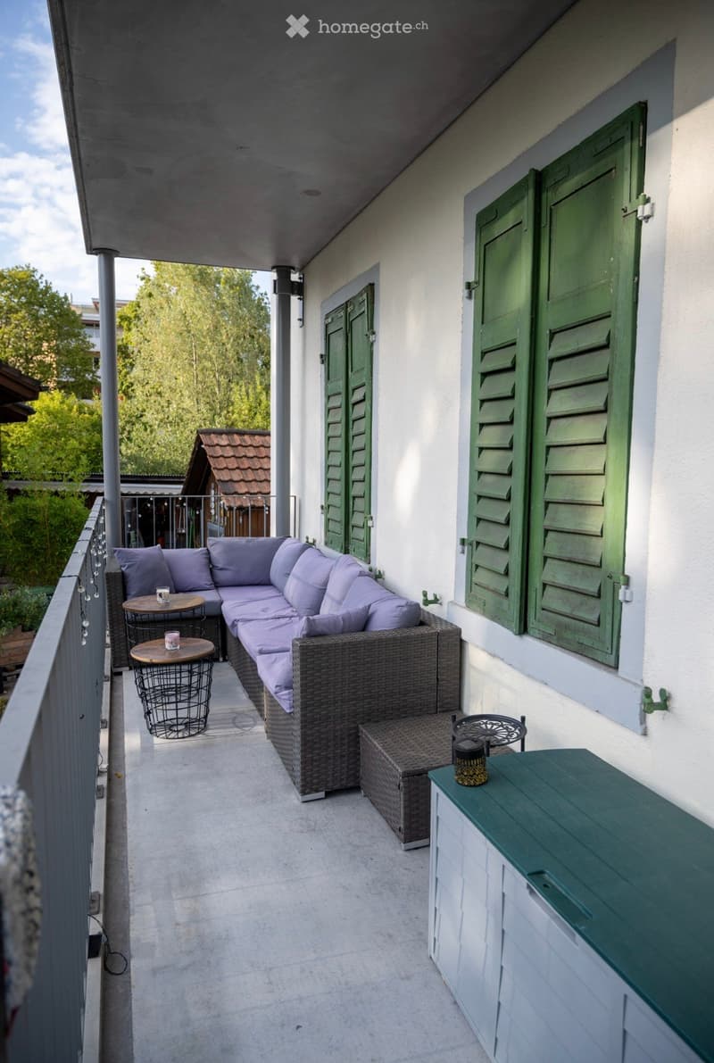 Balcony with a wicker sofa, cushions, small tables, and chairs, overlooking a green area with trees.
