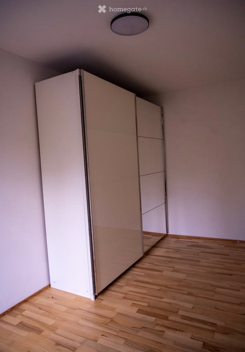 Empty room with light wooden flooring, white walls, and a large mirrored wardrobe unit.