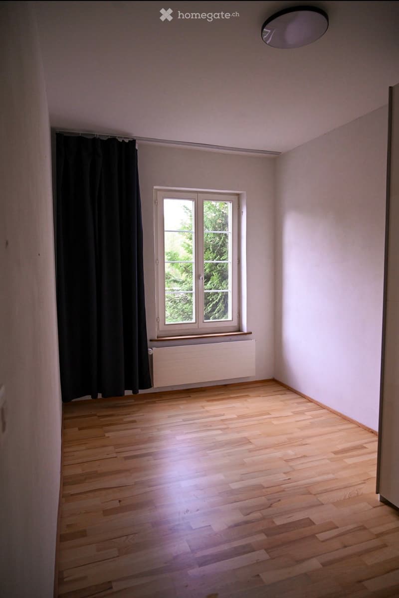 Empty room with wooden flooring, white walls, a window with black curtains, and a radiator below the window.