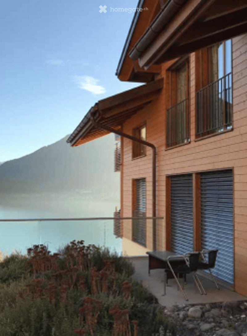 Building. Modern building with large windows and a balcony overlooking a scenic lake and mountain view. Outdoor seating area with a table and chairs is visible.