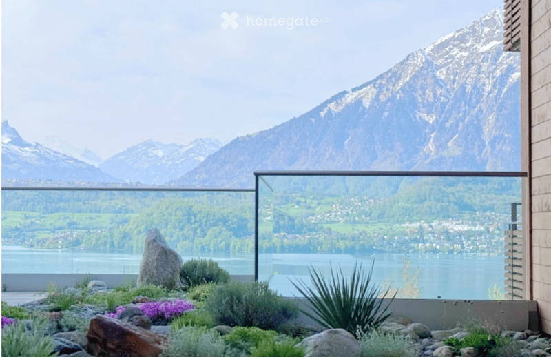 Garden / Outdoor Area. A modern outdoor terrace with a glass railing, overlooking a scenic lake and mountain range. The area features a small garden with rocks, shrubs, and flowers.