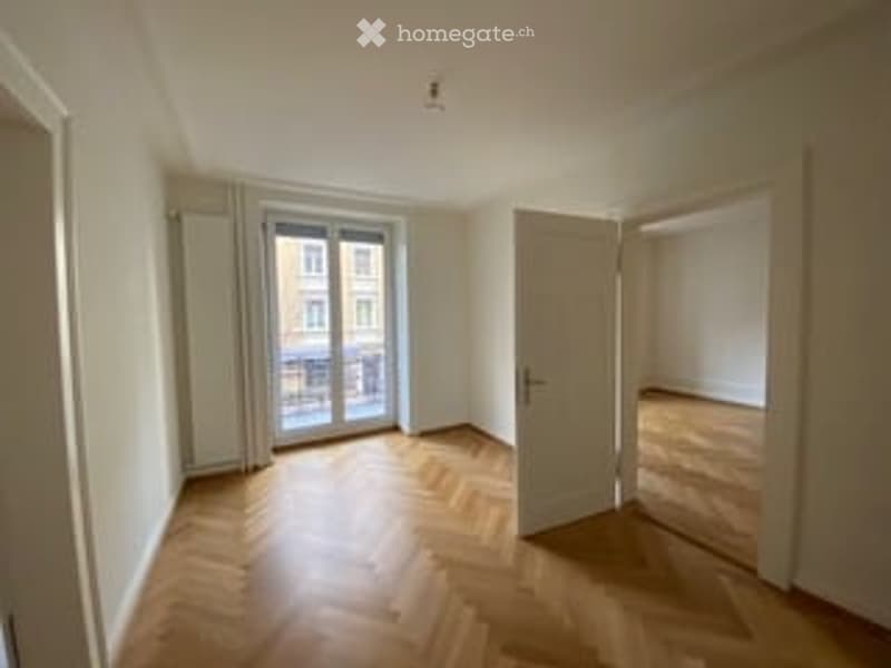 Empty room with wooden flooring, large window with glass door leading to a balcony, and a closed door.