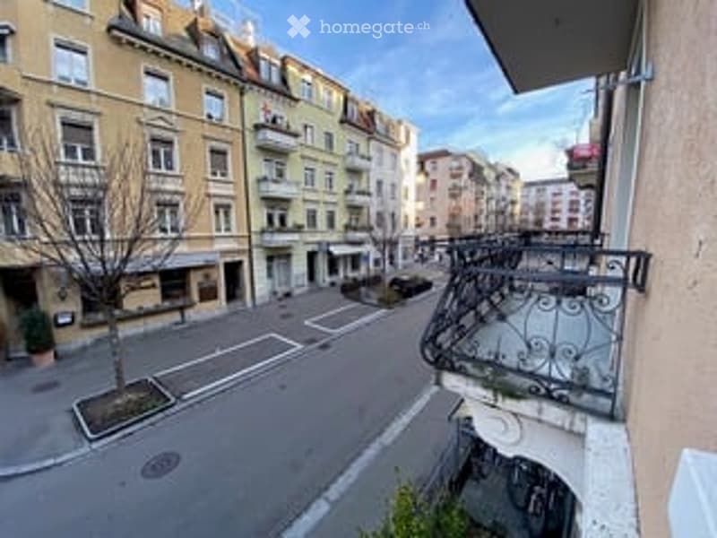 Balcony overlooking a street with buildings, trees, and a parked car visible in the distance.