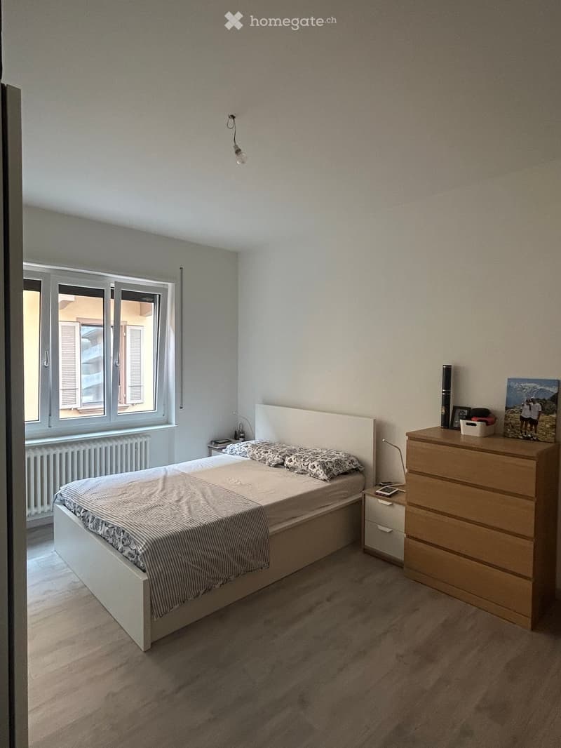 Modern bedroom with a white bed, wooden nightstand, chest of drawers, and a large window with shutters.