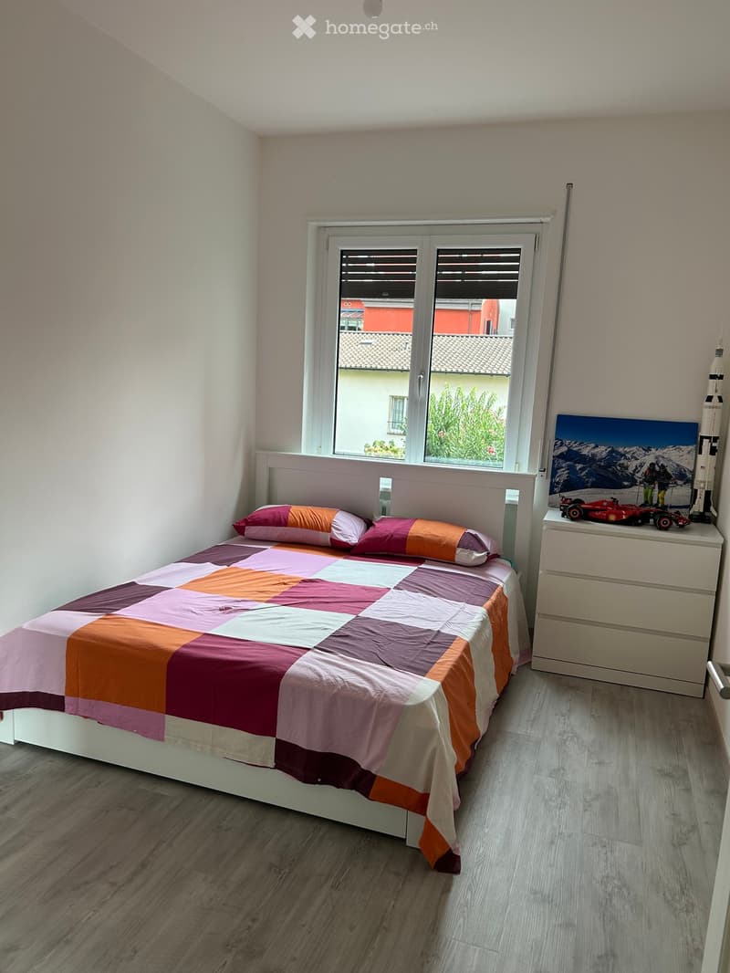 A cozy bedroom with a bed featuring a colorful patchwork quilt and matching pillows, a nightstand with a picture frame, a toy car, and a rocket model, and a window offering a view of a neighboring building and greenery.